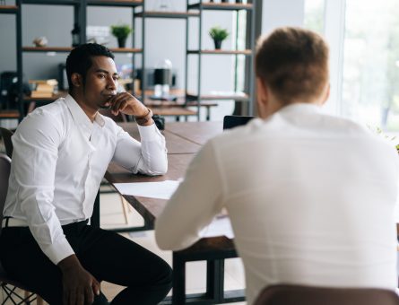 Pensive serious professional African American man interviewing caucasian European young candidate for job at successful company. Two business people talking strategy sitting at office desk.